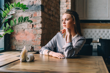 Beautiful young woman sits near big window at cafe, drinks hot tasty beverage, thinks about future plans.