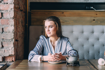 Beautiful young woman sits near big window at cafe, drinks hot tasty beverage, thinks about future plans. Woman rests in coffee bar.