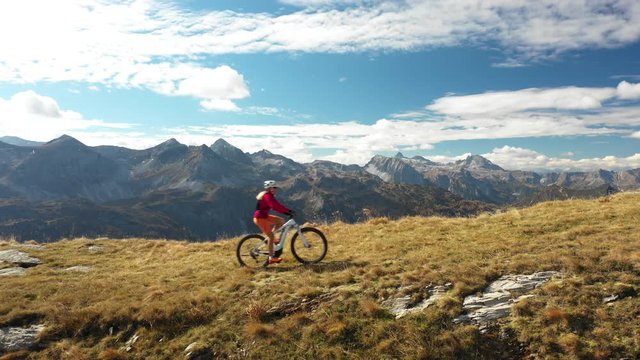 4K sport footage, aerial drone view woman riding electric mountain bike over ridge high up in mountains panorama on sunny autumn day
