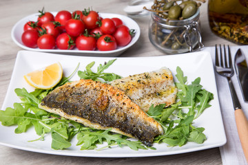 Grilled seabass fillet with arugula, lemon, tomatoes and capers. Close-up.