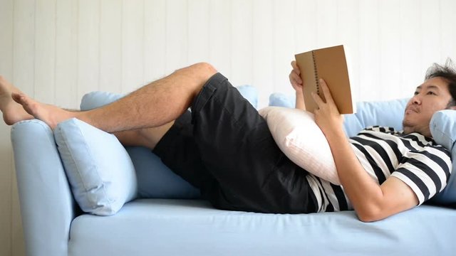 Asian man lying on sofa and reading book at home
