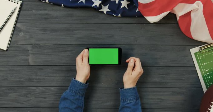 View from above on the black smartphone device lying horizontally on the dark grey wooden desk with rugby ball and American flag while male hands scrolling and taping on the green screen of it. Chroma