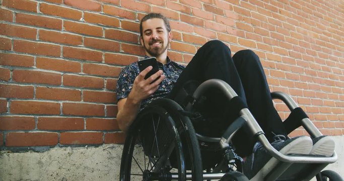 Young disabled man texting in wheelchair leaning against brick wall in city