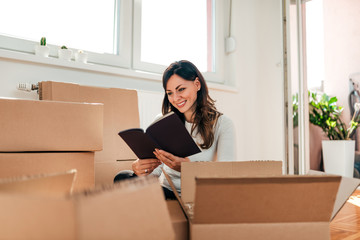 Attractive young woman is moving, reading book and smiling while packing.