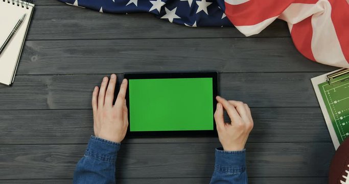 View from above on the black tablet computer lying horizontally on the dark grey wooden desk with rugby ball and American flag while male hands scrolling and taping on the green screen of it. Chroma