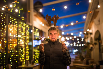 Obraz premium Christmas, people and winter holidays concept - surprised man in horns of deer standing at night street with bengal lights in his hands