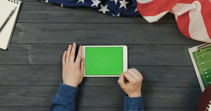 View from above on the white tablet computer lying horizontally on the dark grey wooden desk with rugby ball and American flag while male hands scrolling and taping on the green screen of it. Chroma