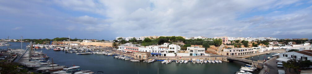 Fototapeta na wymiar A wide panorama view of the town of ciutadella in menorca showing boats moored along the canal and surrounding buildings in bright summer sunlight
