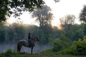 Young woman riding a horse on a beautiful landscape. Clear lake at morning fog.