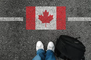 Foto auf Acrylglas Kanada Ein Mann mit Schuhen und Rucksack steht auf Asphalt neben der Flagge Kanadas und der Grenze