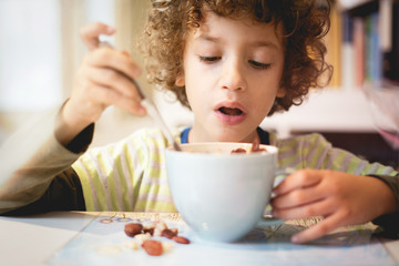 Cute boy with curly hairs making his breakfast eating milk and chocolate muesli