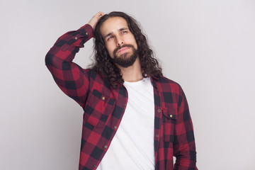 confused handsome man with beard and black long curly hair in casual checkered red shirt standing, thinking and looking away with thoughtful face . indoor studio shot, isolated on grey background.