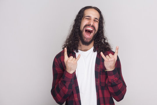 Rock and roll. funny man with beard and black long curly hair in casual checkered red shirt looking ar camera with rock sign gesture and screaming. indoor studio shot, isolated on grey background.