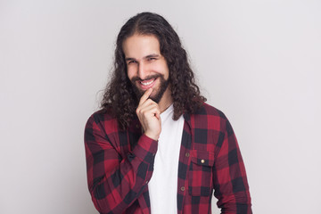 Portrait of playful handsome man with beard and black long curly hair in casual style, checkered red shirt standing looking at camera with cunning face. indoor studio shot, isolated on grey background