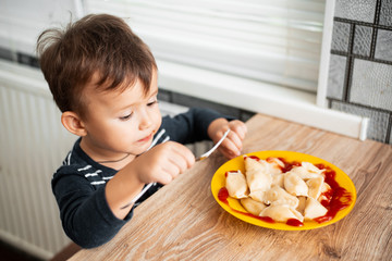 Hungry child eating dumplings in the kitchen, sitting at the table in a gray jacket
