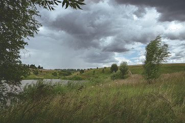 Cloudy windy summer landscape with small river.Dark stormy clouds in dramatic overcast sky.Fields,green meadows and woods.Rainy season. 