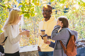 Enjoying student life in sunny autumn day. Smiling two young women and black man with coffee on...