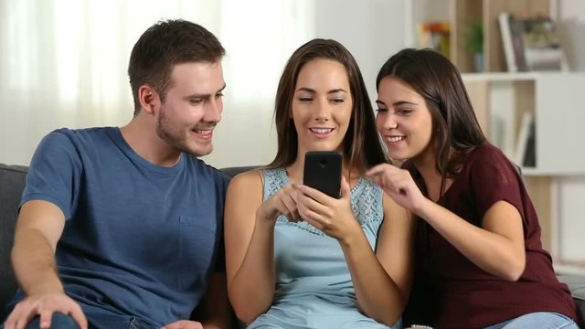 Front view of three happy friends talking about smart phone content at home sitting on a couch
