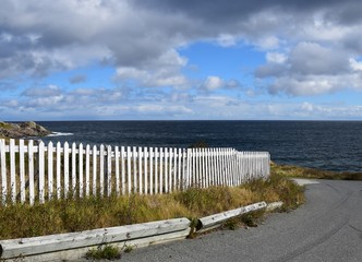 road leading to a blue ocean  lined with a white picket fence with blue ocean and blue cloudy sky 