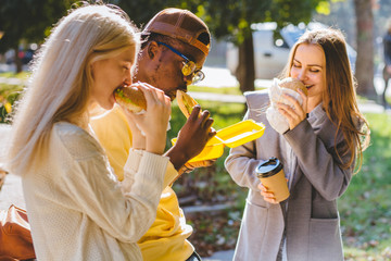 Three young multiethnic hungry people students discussing, having lunch, and eating to-go food while standing up outdoor in sunny autumn day. Making friends, get-together meeting concept.