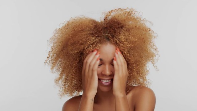 mixed race woman with big curly afro blonde hair hide her eyes behind her hands and then show it up and smiling and dancing