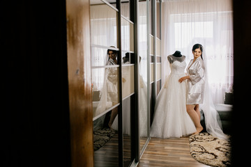 The charming bride with bridesmaids stands near wedding dress. Bridal wedding morning preparation. 