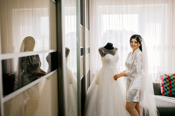 The charming bride with bridesmaids stands near wedding dress. Bridal wedding morning preparation. 