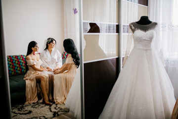 The charming bride with bridesmaids stands near wedding dress. Bridal wedding morning preparation. 