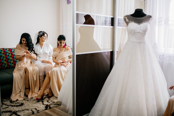 The charming bride with bridesmaids stands near wedding dress. Bridal wedding morning preparation. 