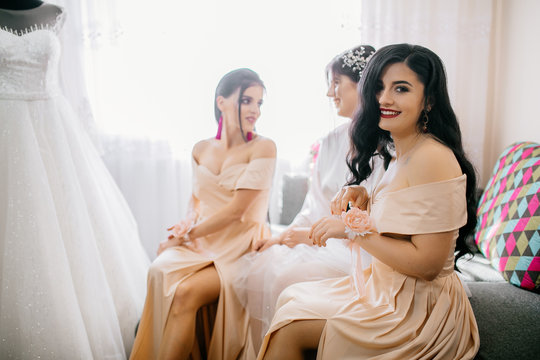 Bridesmaids helping slender bride lacing her wedding white dress, buttoning on delicate lace pattern with fluffy skirt on waist. Morning bridal preparation details newlyweds. Wedding day moments.