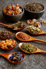 Spoons with different types of dry tea leaves on rusty dark background. Top view.