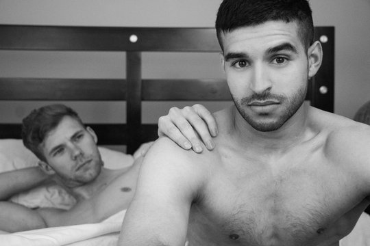 Gay lovers sitting in bed, one of them looks at camera as other looks at his partner