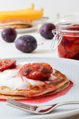 White wooden table with closeup image of dessert saucer with crumpets, pancakes, sour cream and fresh plums. The glass with cooked plums, fresh plums and fresh bananas in the background.