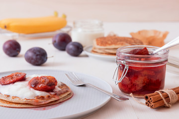 White wooden table with closeup image of dessert saucer with crumpets, pancakes, sour cream and fresh plums. The glass with cooked plums, fresh plums and fresh bananas in the background.