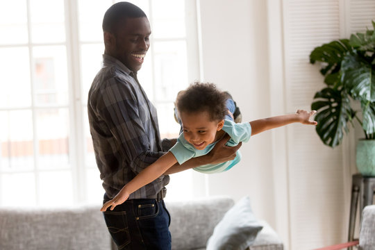 African American Family Spend Time Together Have A Fun Standing In Living Room At Home. Laughing Father Holding On Hands Flying Smiling Cute Little Preschool Toddler Son. Happy Black Family Concept