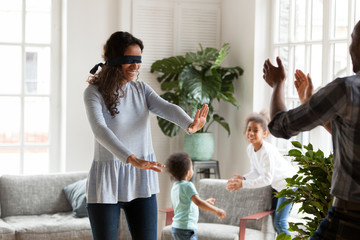 Happy black funny family with little preschool son and daughter spending free time together on weekend at new modern home. Playful mom with covered eyes playing blindman hide and seek in living room