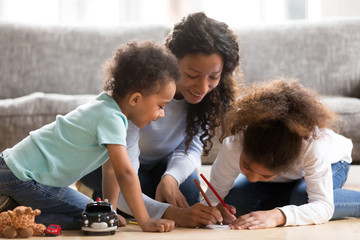 Black African loving mother help to their children draw. Positive mom sitting with toddler son and preschool daughter on a wooden warm floor spend free time on weekend together in living room at home