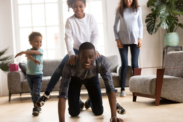 Fototapeta premium Black African wellbeing attractive family spend free time on weekend have a fun together in living room at new modern home. American happy laughing father rolls little preschool daughter on his back