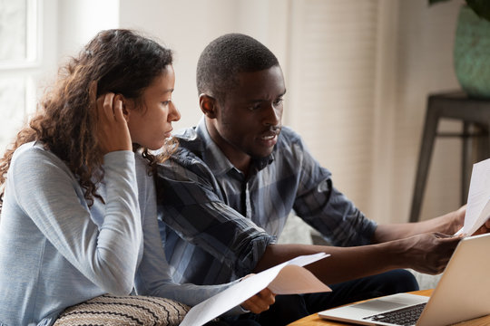 African Millennial Stressed Married Couple Sitting On Sofa At Home Checking Unpaid Bills, Taxes, Due Debt, Bank Account Balance. Bankruptcy, Debt And Lack Of Money Financial Problems In Family Concept