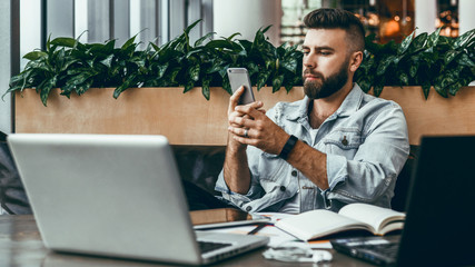 Hipster man sits in cafe, uses smartphone, works on two laptops. Businessman reads an information message in phone.