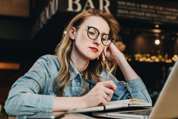 Sad student girl sitting in cafe in front of laptop preparing for exams. Woman takes boring online...