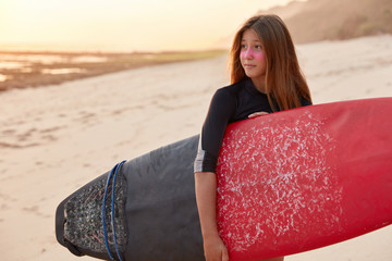 Shot of surfer woman in black swimsuit, stands against ocean horizon background, holds surfboard, has stroll across sea or ocean, waits waves, finds perfect sport, admires sunshine. Recreation concept