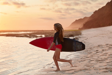 Fototapeta na wymiar People and travel concept. Shot of slim surfer with perfect figure, being in good shape as involved in active summer activities, holds red surfboard in hand, goes in direction to ocean waits for waves