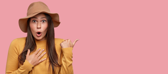 This knocks me from feet. Excited brunette young woman has bated breath, points with thumb, wears beige hat and yellow sweater, isolated over pink background with free space for your promotion