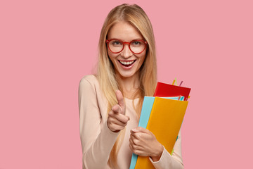 Joyful blonde female model wears round spectacles, makes gun gesture, holds textbooks, has toothy...