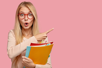Fototapeta Photo of surprised teacher indicates aside with index finger, has light straight hair, keeps mouth opened with amazement, carries textbooks, isolated on pink background. Omg, look at this space obraz