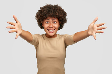 Happy satisfied dark skinned girl gives hug at camera, spreads hands, meets with close friend, dressed casually, isolated over white background. Come to me, I want to embrace you. Pleasant feelings