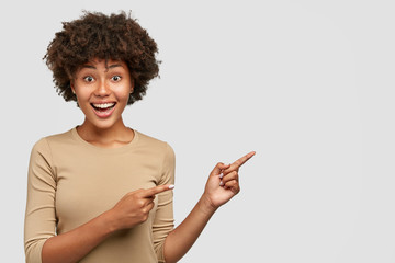 Indoor view of cheerful emotive ethnic young woman with satisfied expression, indicates aside at...