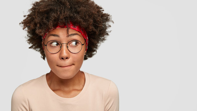 Headshot of puzzled mixed race woman presses lips and look with bewilderment aside, notices something strange, has Afro bushy hairstyle, dressed in casual clothes, isolated on white background