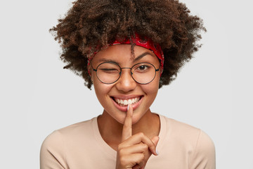 Headshot of playful cheerful young Afro woman makes shush gesture with positive expression, blinks...